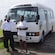 A man and a woman pose in front of a minibus with passengers on it